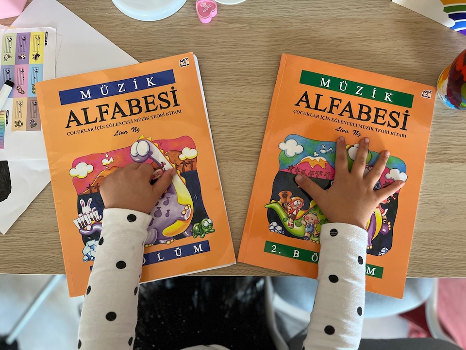 Maya's hands resting on two Müzik Alfabesi books by Lina Ng — Volume 1 completed, Volume 2 just starting